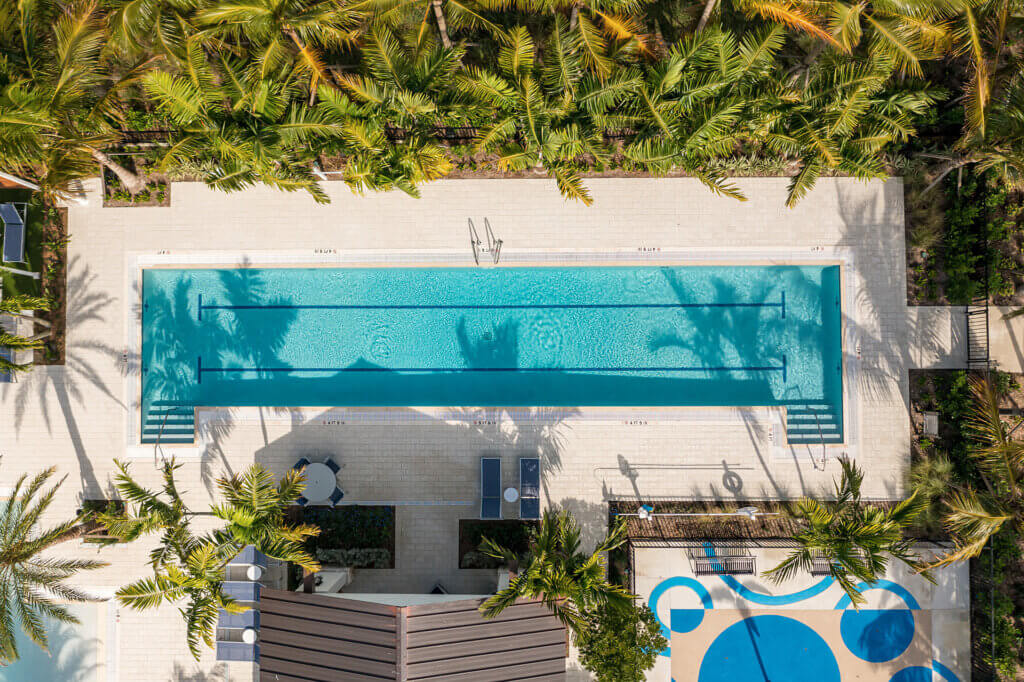 Aerial view of a rectangular swimming pool at Catalina Palms at Sundance, surrounded by palm trees and lounge chairs. Ladders stand at each end, and a blue and tan circular pattern decorates the nearby area. Sunlight casts shadows across the scene.