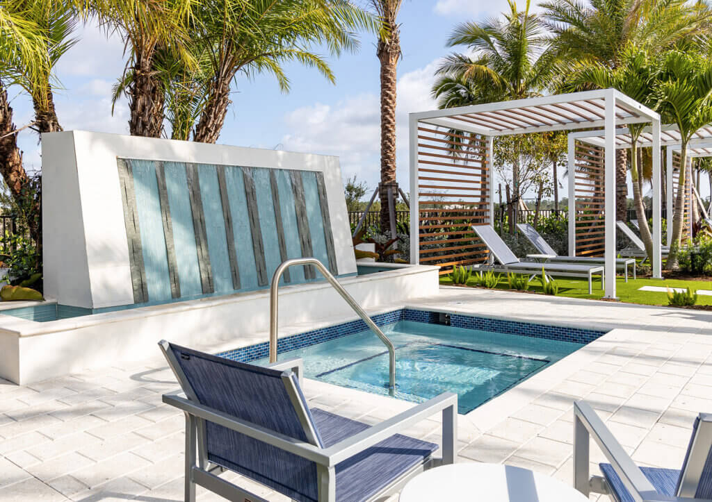 The Catalina Palms at Sundance boasts a modern outdoor pool area with a small square spa, a water feature, and a shaded lounge. Surrounded by tall palm trees, lounge chairs, and under a sunny sky, the light-colored tile flooring adds to its serene charm.
