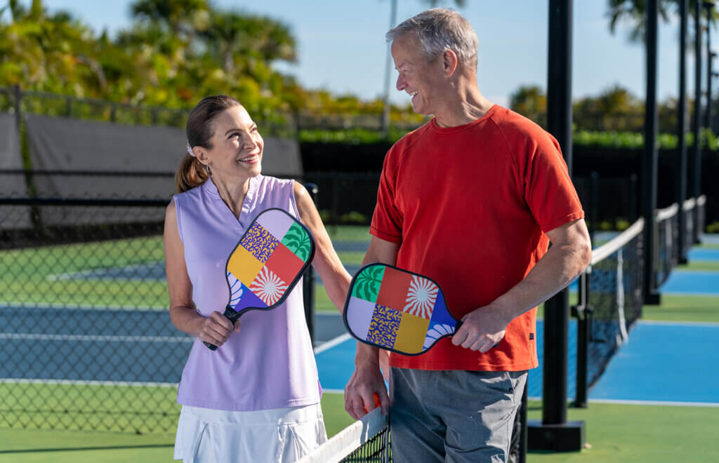 A man and a woman are standing on a pickleball court holding paddles. The woman is wearing a light purple top and white skirt, and the man is in a red shirt and gray shorts. They are smiling at each other. Palm trees are visible in the background.