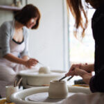 Two people are working on pottery at a studio. The foreground shows a person using a tool on a clay pot on a pottery wheel. In the background, another person is focused on their own pottery wheel. The setting is well-lit with natural light.