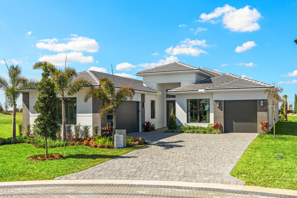 Single-story modern house with attached two-car garage, landscaped yard, and paved driveway on a sunny day. Streetlamp and green lawn in foreground.