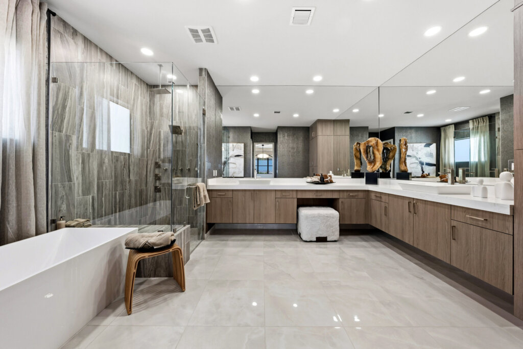 Modern bathroom with large glass shower, freestanding tub, double vanity with wood cabinets, a mirrored wall, and tile flooring. Decorative accents and a stool are present.