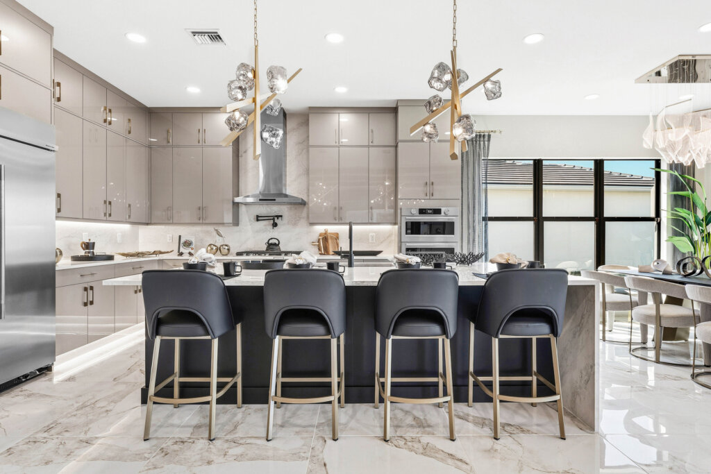 Modern kitchen with glossy cabinetry, marble floors, a central island with four dark barstools, pendant lights, stainless steel appliances, and an adjacent dining area with large windows.