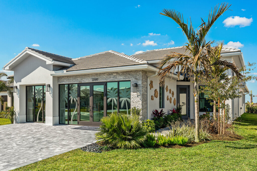 Single-story modern house with large glass windows, light-colored exterior, tiled roof, paved driveway, green lawn, and palm trees under a blue sky.