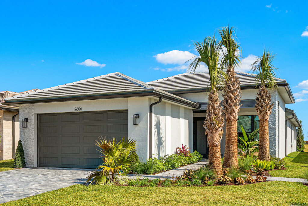 Single-story modern house with a two-car garage, palm trees, and well-maintained lawn under a clear blue sky.