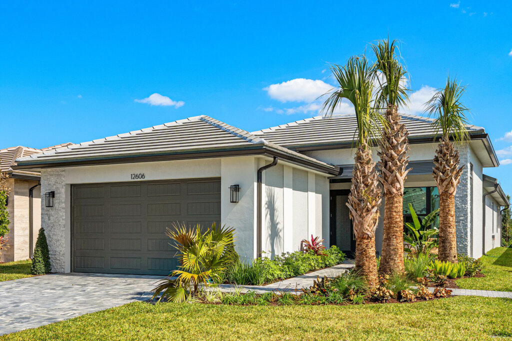 Single-story modern house with a two-car garage, palm trees, and a neatly landscaped front yard under a clear blue sky.