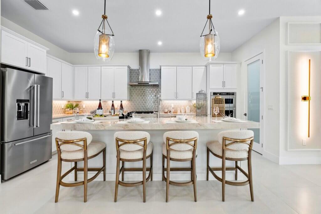 A modern kitchen with white cabinetry and a large island featuring four wooden chairs with cushioned seats. Pendant lights hang above the island, and stainless steel appliances, including a refrigerator and an oven, are visible.