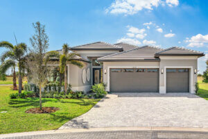 Modern single-story house with a three-car garage, palm trees, and a paved driveway, set on a grassy lot under a blue sky with scattered clouds.