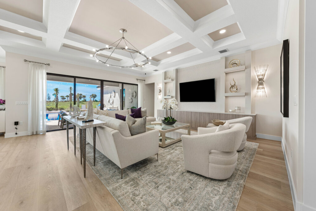 Modern living room with neutral decor, two armchairs, a sofa, a large TV, built-in shelves, ceiling lights, and sliding glass doors opening to a pool and outdoor area.