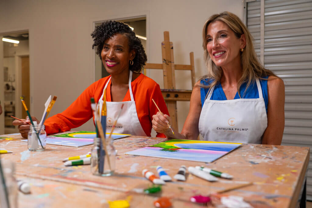 Two women wearing aprons sit at a table in an art studio, smiling as they paint on canvases with brushes and paint tubes scattered on the table.