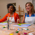 Two women wearing aprons sit at a table in an art studio, smiling as they paint on canvases with brushes and paint tubes scattered on the table.