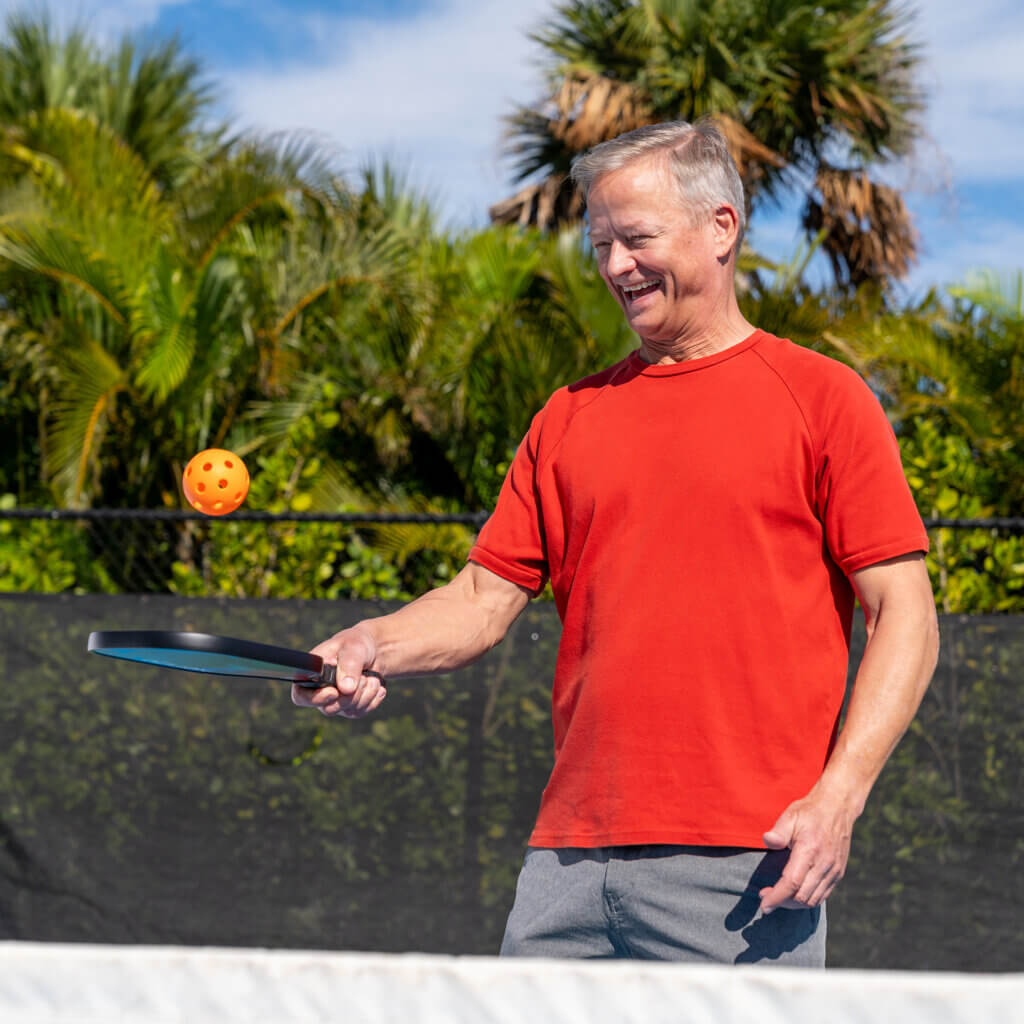 A man in a red shirt is playing pickleball outdoors. He is holding a paddle and about to hit an orange ball. There are green palm trees and a blue sky in the background.