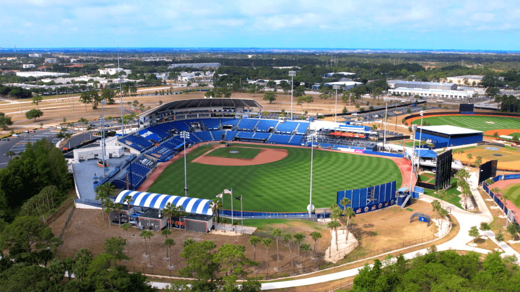 Aerial view of a baseball stadium with a green field and surrounding blue seats. The stadium is set in a landscaped area with trees and pathways. Nearby are smaller fields and parking lots. The sky is partly cloudy.