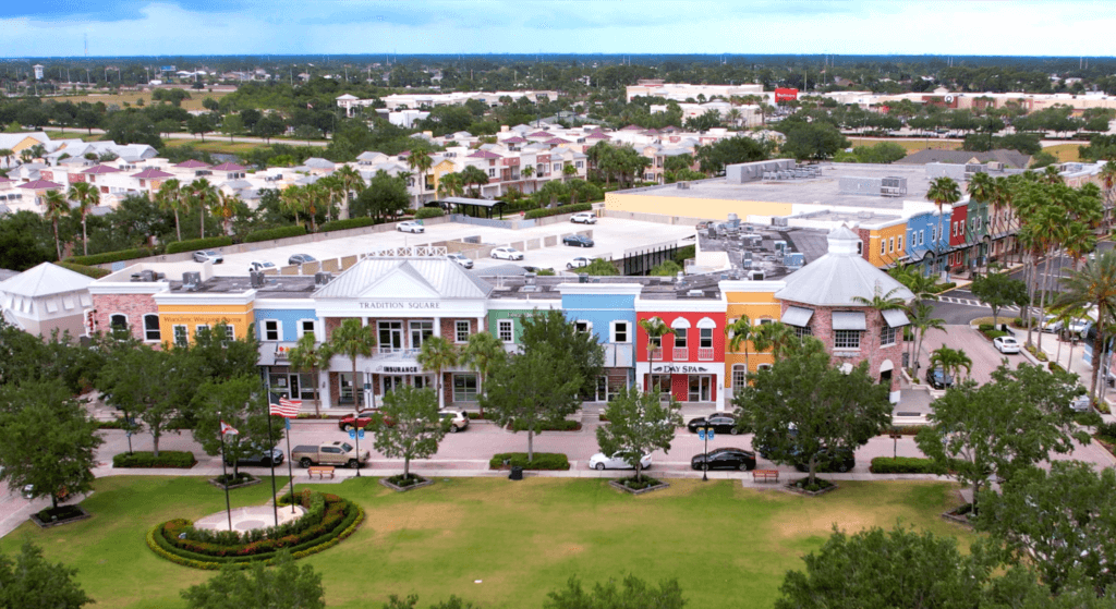 Aerial view of a town square with a grassy lawn surrounded by trees in the foreground. Colorful buildings line the perimeter, and several cars are parked along the streets. A flagpole stands in the grass. Residential areas are visible in the background.