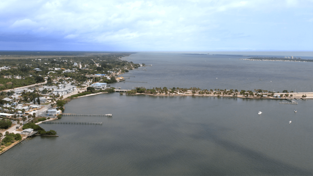 Aerial view of a coastal town with a large body of water extending into the distance. The shoreline features buildings, docks, and greenery. Boats are visible on the water, and clouds are scattered across the sky.