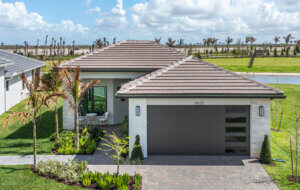 Single-story modern house with a two-car garage, manicured lawn, and stone driveway, set in a suburban neighborhood with a pond and palm trees in the background.