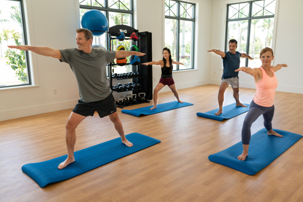 Four adults practice yoga on blue mats in a bright room, all holding a standing pose with arms extended, facing forward.