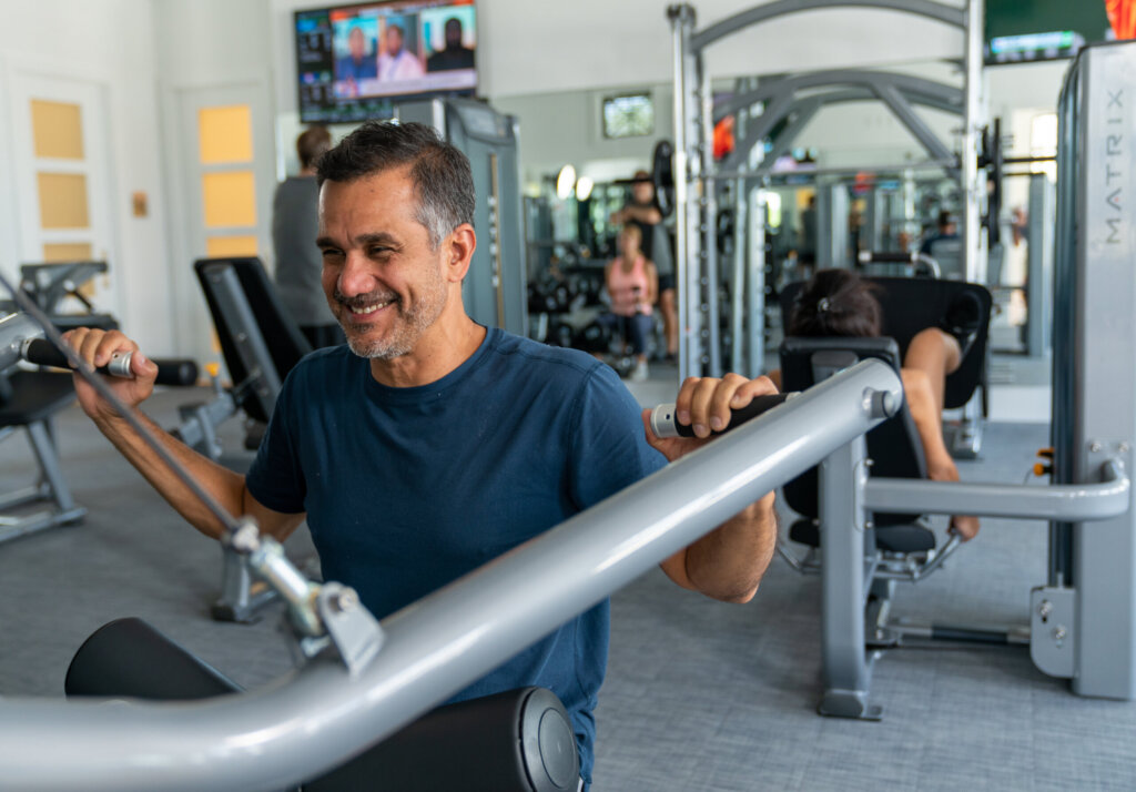 Man in blue shirt using a machine for seated shoulder presses at a gym; other people and exercise equipment are visible in the background.