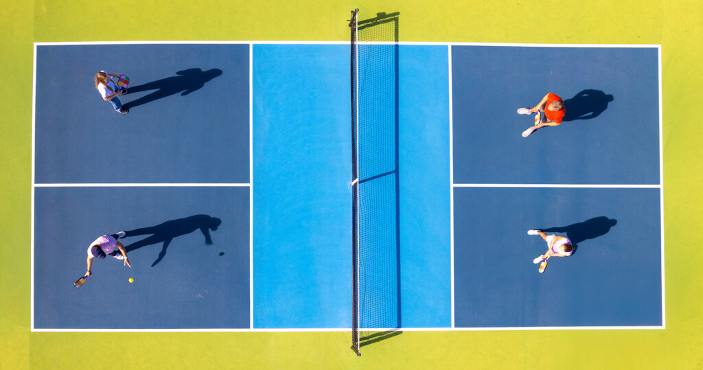Aerial view of four people playing doubles pickleball on a blue and green outdoor court, with the ball in play near one player.
