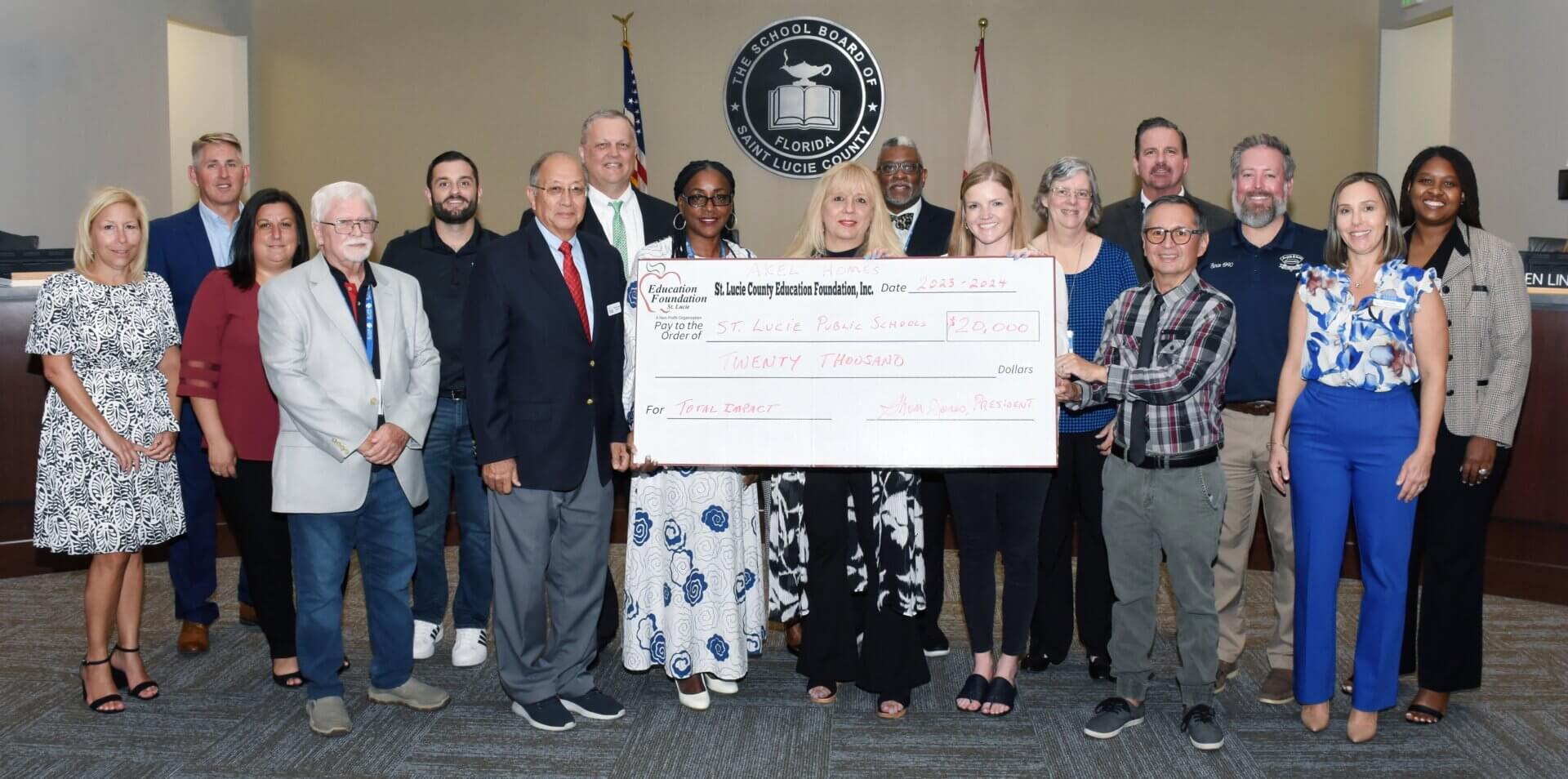 A group of people stands indoors, holding a large ceremonial check presented by the St. Lucie County Education Foundation to St. Lucie Public Schools for $20,000 to support Student Innovation. Thirteen men and women pose together, with a table and American flag in the background.