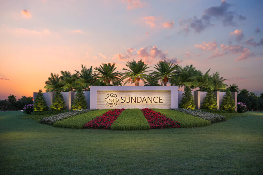 A landscaped area features a large sign with the word "Sundance" in the center. The sign is surrounded by palm trees and neatly arranged plants, including red and green foliage. The sky is partly cloudy with a warm sunset glow.